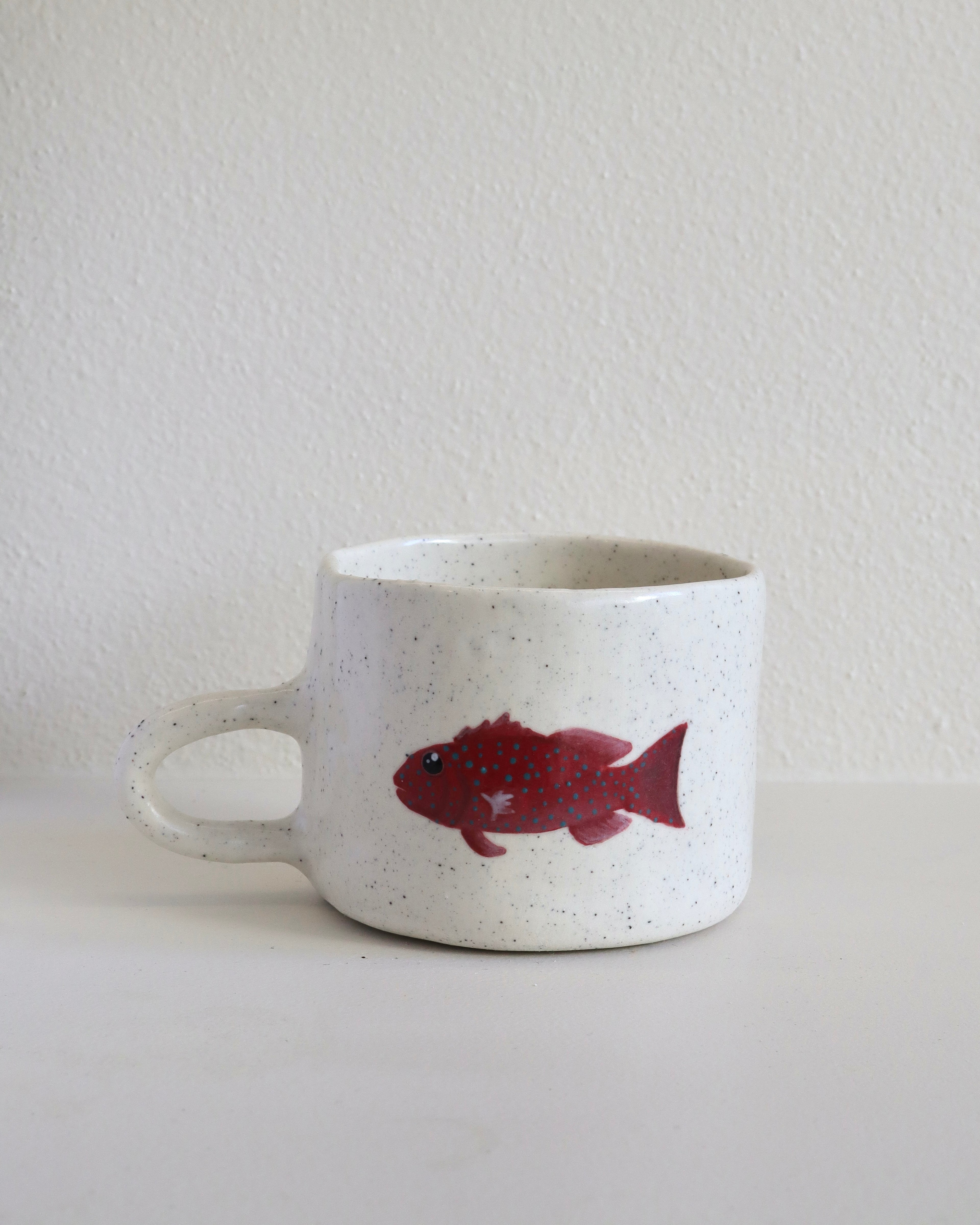 A speckled white ceramic mug with a red fish illustration on the side, set against a plain white wall and surface.