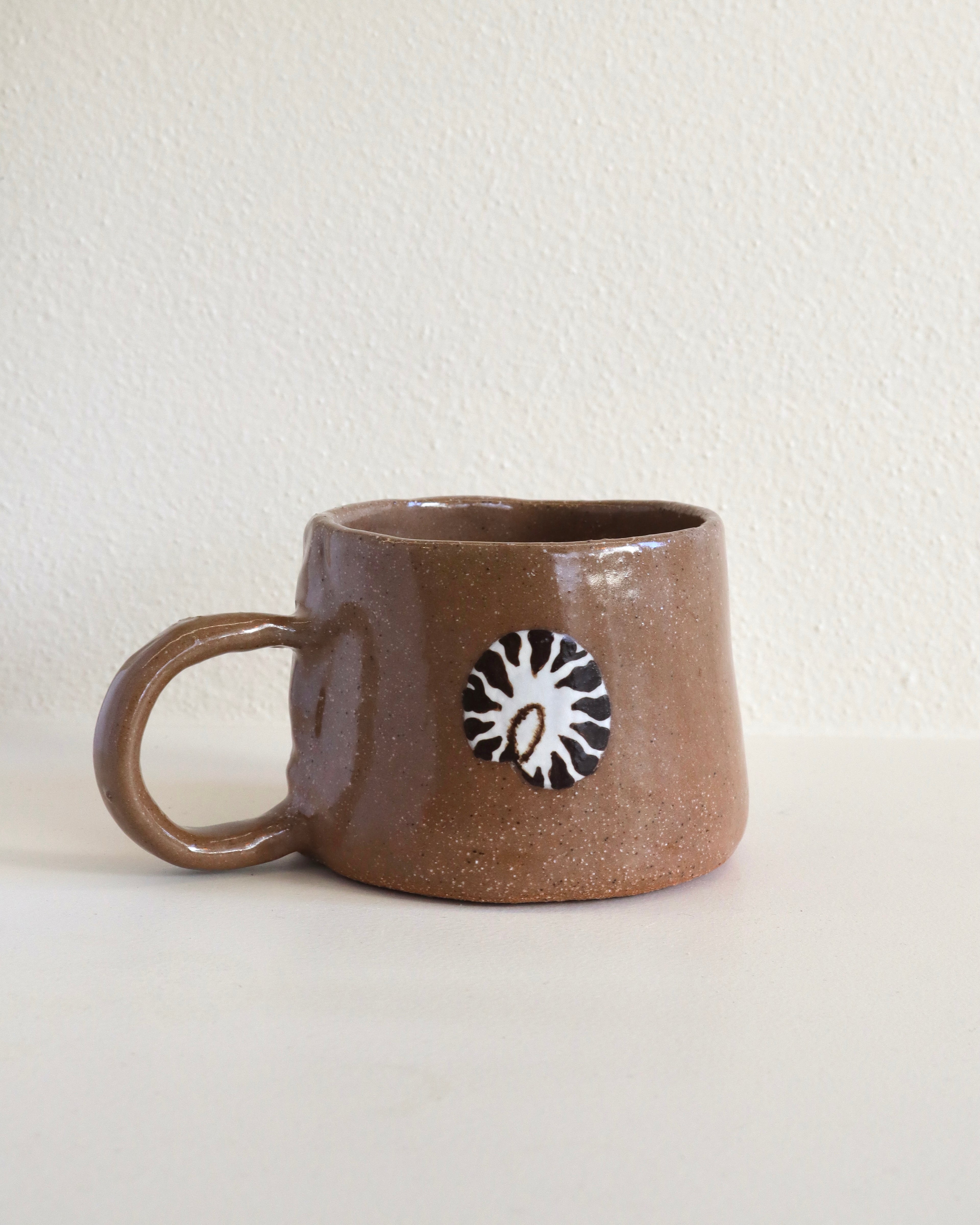 A handmade brown ceramic mug with a textured glaze and a nautilus hand painted design on the front, set against a plain off-white wall.