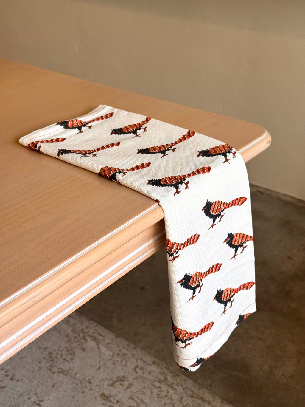 100% cotton block printed teatowel featuring australian bird Pheasant Coucal. Striking rust orange and black feathers are the signature mark of the coucal. draped across a table. 