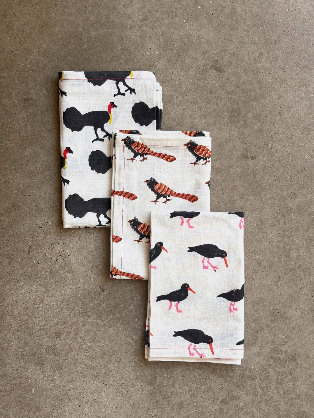 Three folded white cotton kitchen tea towels with hand block printed bird prints are arranged on a textured gray concrete surface. The towels feature black birds, brush turkeys, red-and-black striped pheasant councils, and pink-and-black sooty oyster catcher birds. All native australian birds.