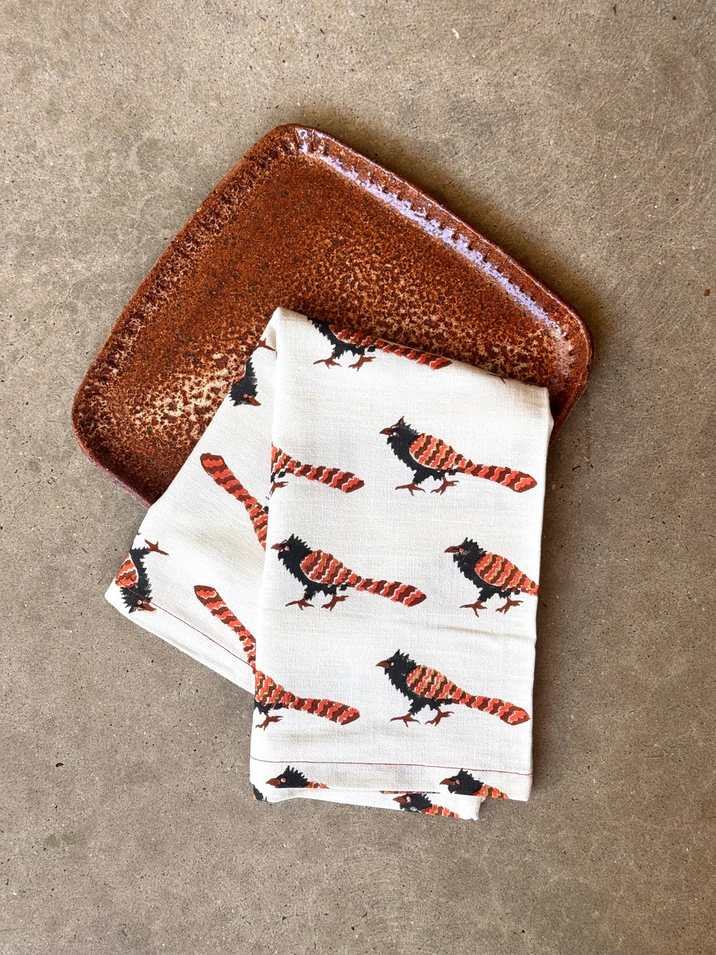 A folded white cotton tea towel with a repeating pattern of black and orange striped pheasant coucal birds, displayed on a textured brown ceramic plate over a neutral concrete surface. Block printed design on 100% cotton. 