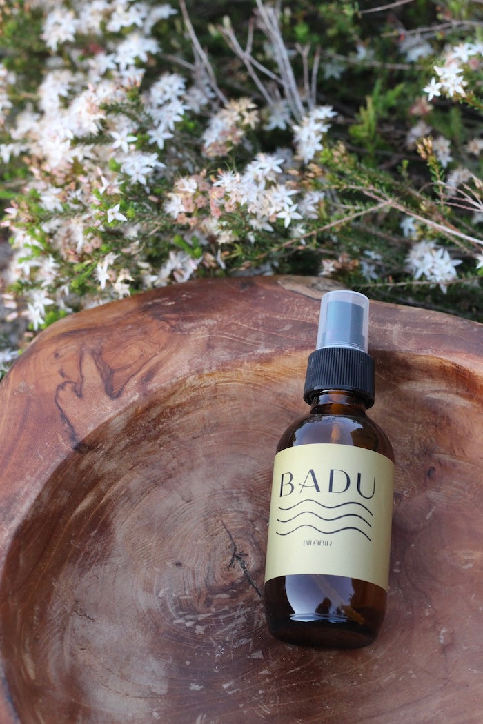 Bottle of BADU Bilabir aromatherapy mist on a wooden surface with a natural nativie floral background