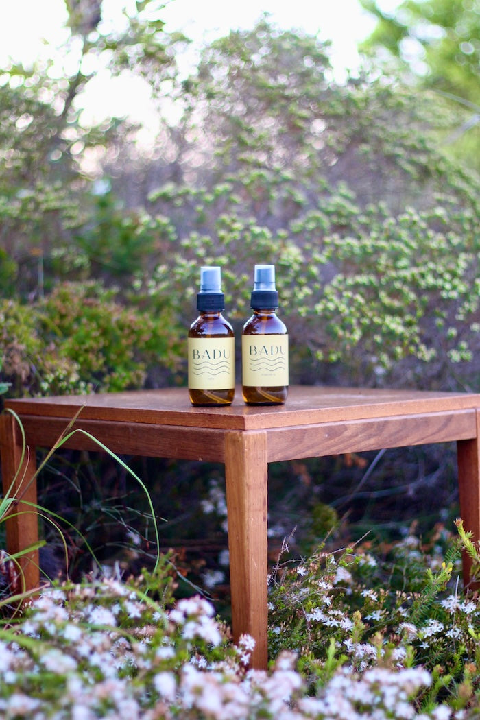 Two bottles of Badu Natural Aromatherapy Mist on a wooden table with a natural Native floral background