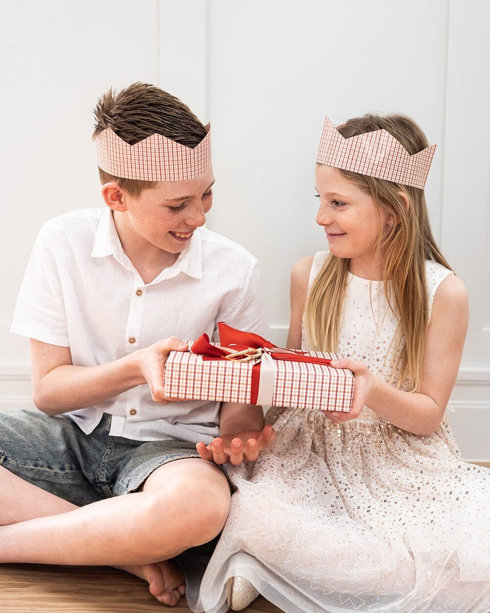 Two children wearing Enchanted red check paper crown hats while opening a gift