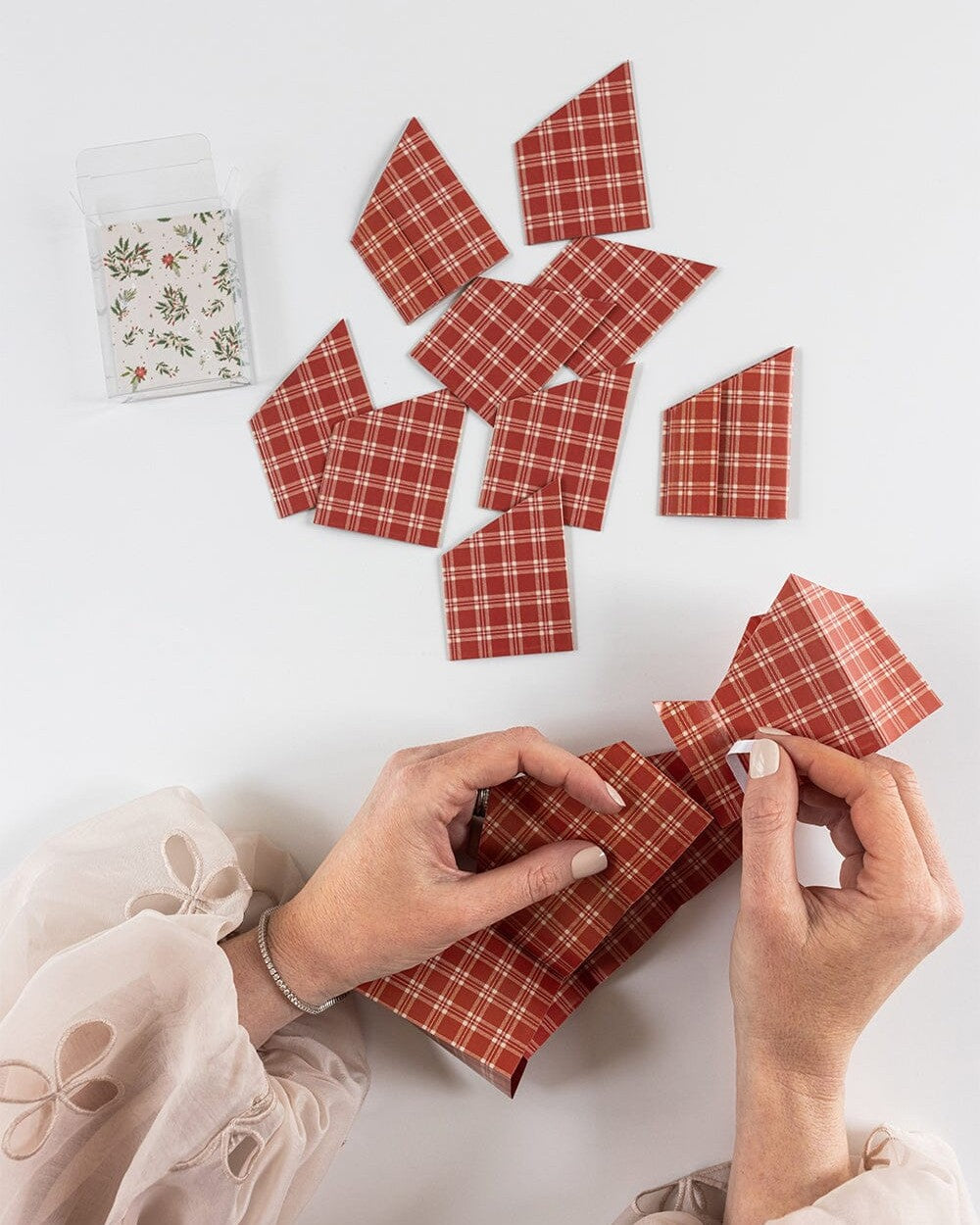 Hands folding Heirloom Holly red plaid paper crowns beside the box