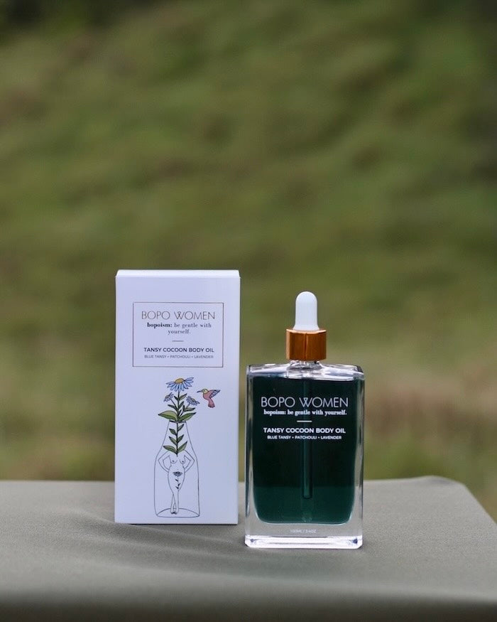 Bopo Women's Tansy Cocoon body oil bottle and packaging on a blurred green background